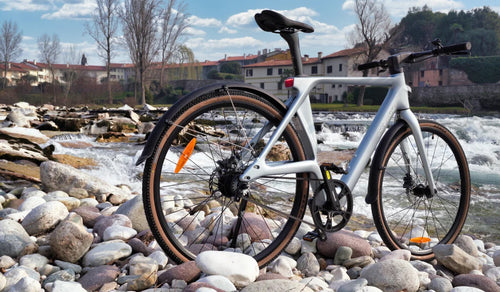 Fiido Air ultra-lightweight carbon-fibre electric bike displayed on a rocky riverbank beside flowing water.