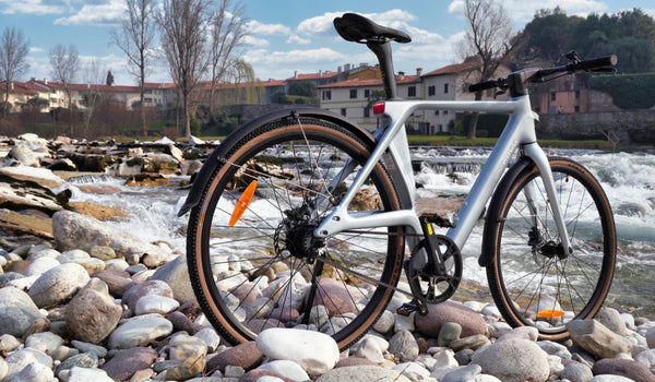 Fiido Air ultra-lightweight carbon-fibre electric bike displayed on a rocky riverbank beside flowing water.