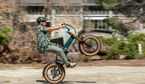 A rider performs a wheelie on a Fiido M1 Pro fat tire e-bike, showcasing its compact folding frame, wide tires and front suspension on an urban road.