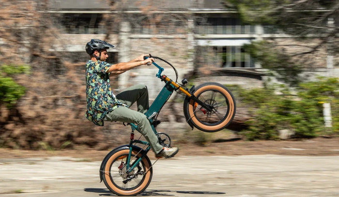 A rider performs a wheelie on a Fiido M1 Pro fat tire e-bike, showcasing its compact folding frame, wide tires and front suspension on an urban road.